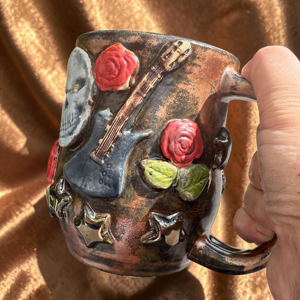 Rock N Art Shop handmade stoneware mug featuring sculpted skulls, guitars, red roses, and stars with a bronze glaze.