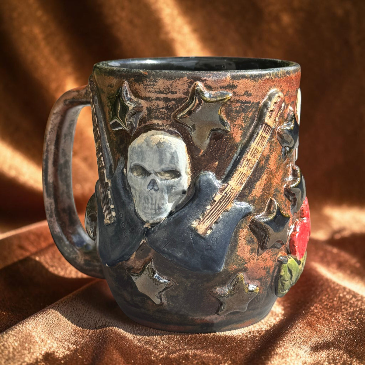 Rock N Art Shop handmade stoneware mug featuring a sculpted skull, electric guitars, and stars in a bronze glaze.