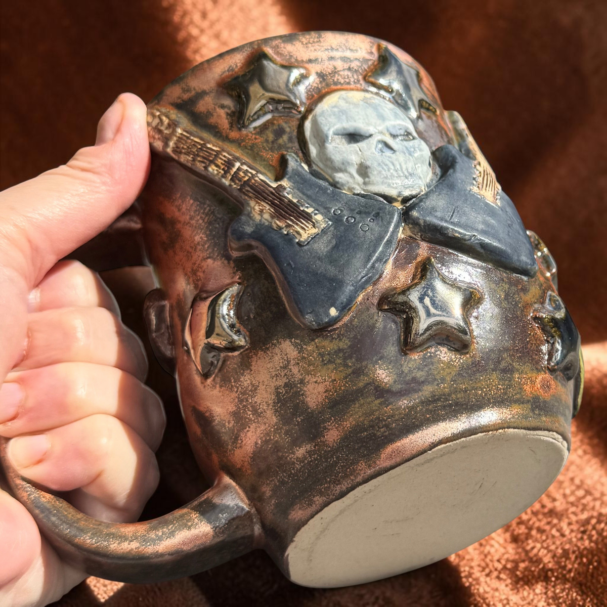 Rock N Art Shop handmade stoneware mug featuring sculpted skulls, electric guitars, and stars with a bronze glaze.