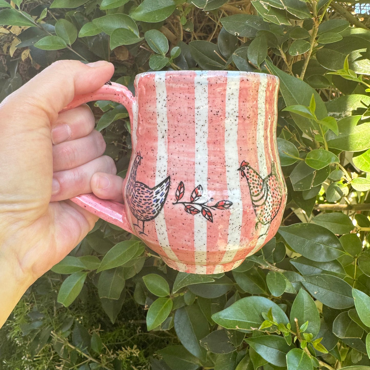 Hand holding a pink striped mug with bird designs against a green leafy background