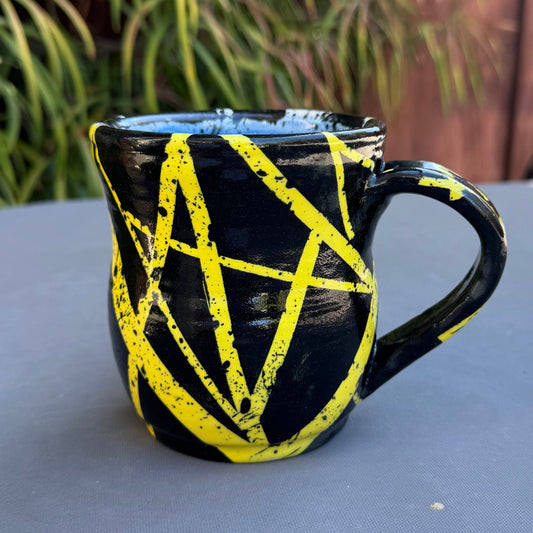 A black ceramic mug with a handle, decorated with bright yellow painted intersecting lines, sitting on a gray table outdoors with a wooden fence and green plants in the background.