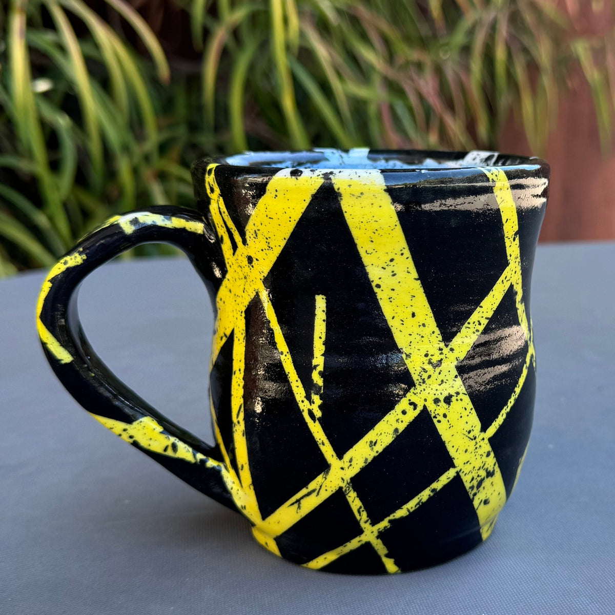 A black ceramic mug with a bold yellow painted crisscross pattern, sitting on a gray table outdoors with a wooden fence and green plants in the background.