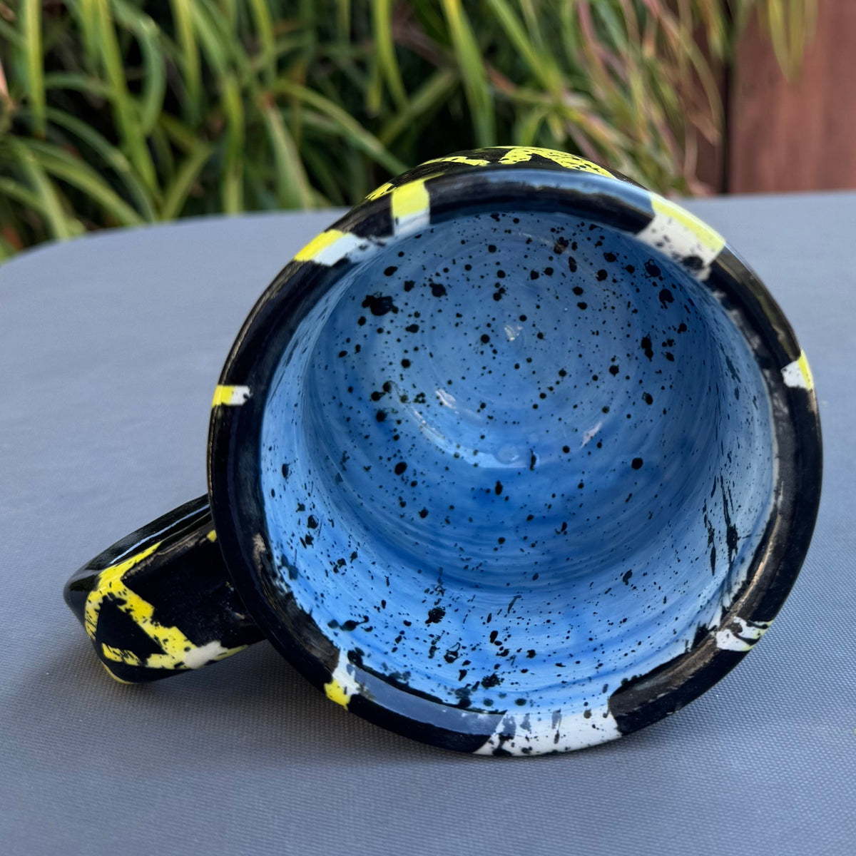 A handmade ceramic mug with a blue speckled interior and a black exterior decorated with yellow and white accents, resting on a gray table with a wooden fence and green plants in the background.