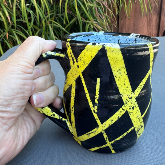 A hand holding a black ceramic mug with bold yellow painted diagonal lines, set against an outdoor background with green plants and a wooden fence.
