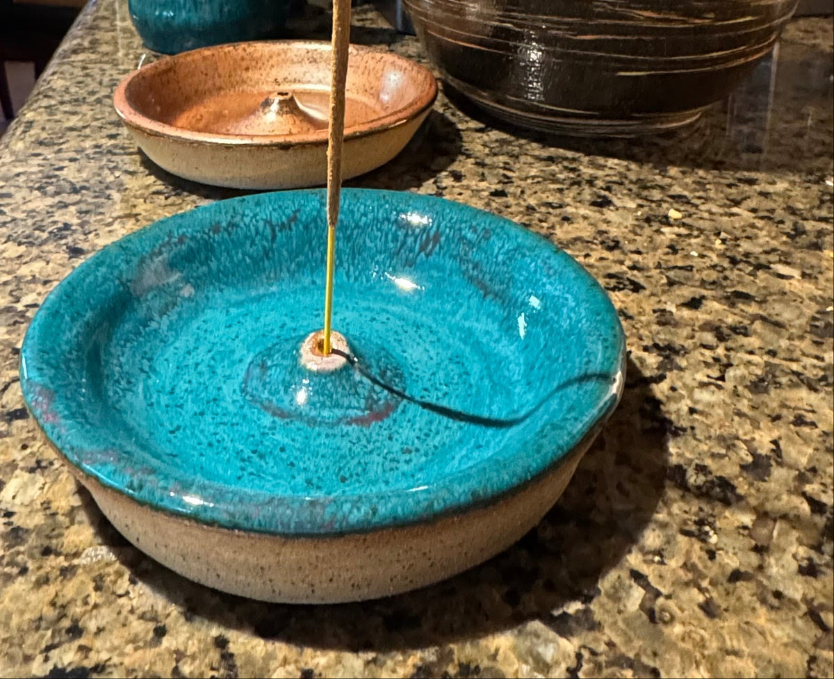 A handmade ceramic incense burner in a vibrant turquoise glaze with a speckled beige base, shown on a granite countertop with other ceramic dishes in the background.