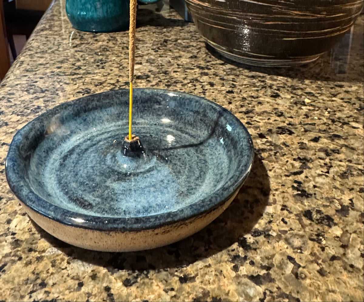 A handmade ceramic incense burner dish with a blue-gray glaze and a beige base, shown on a granite countertop with an incense stick inserted. Other ceramic items are visible in the background of a home kitchen setting.