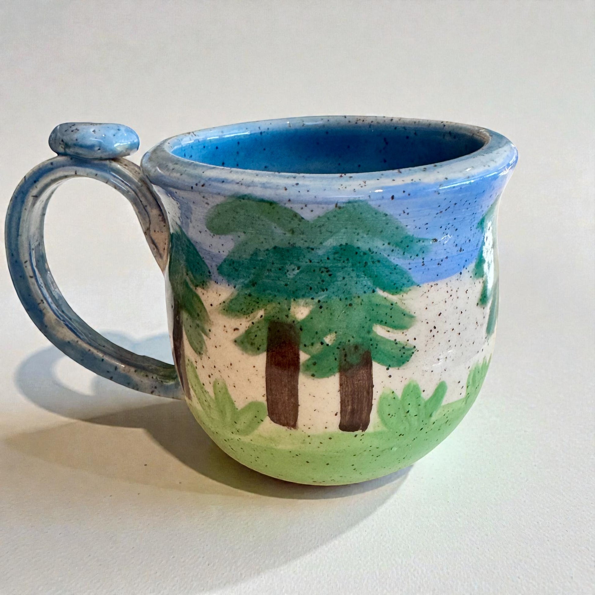 Rock N Art Shop handmade stoneware mug with raised forest scene, trees, and blue sky glaze. 12 oz capacity.
