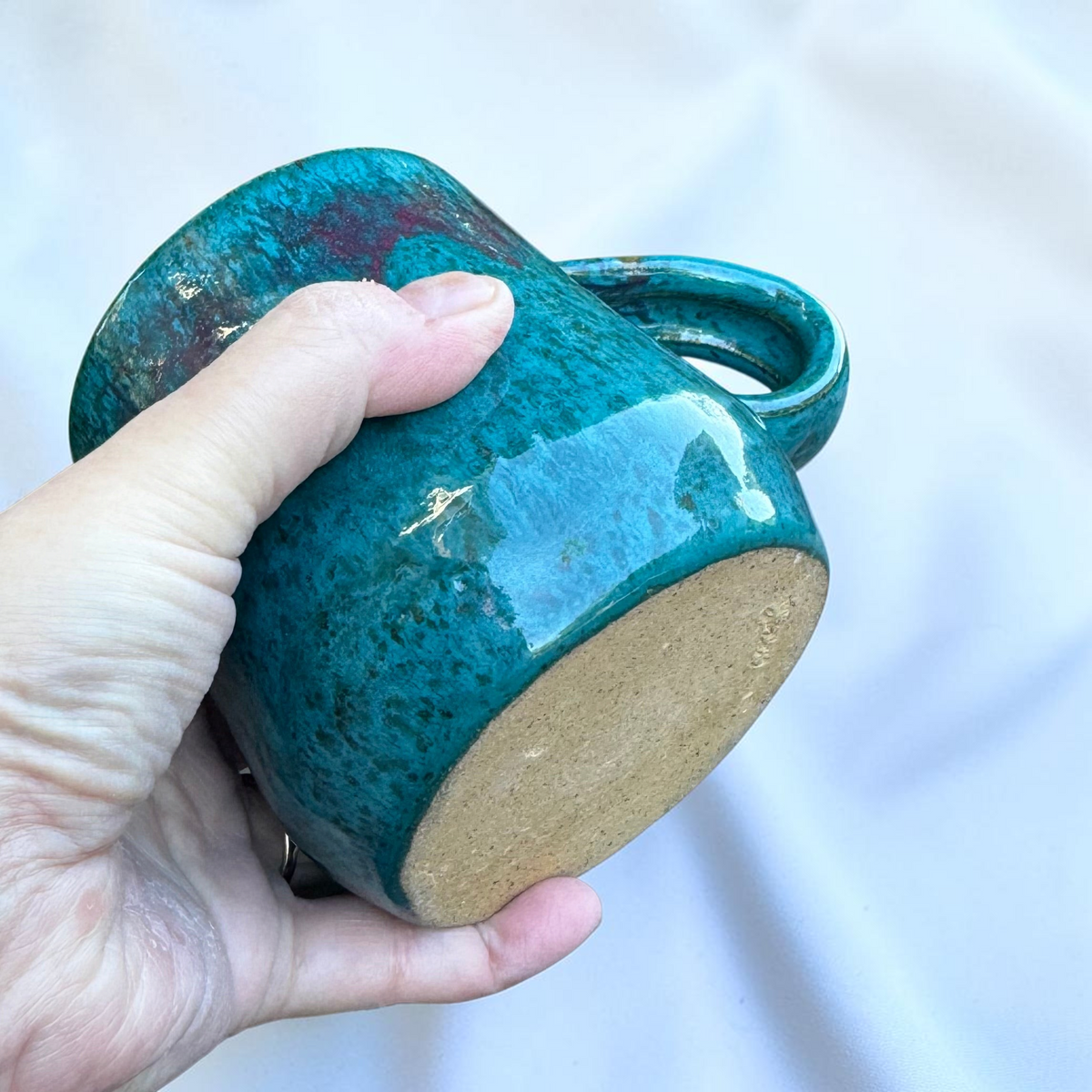 Rock N Art Shop handmade speckled stoneware coffee mug in glossy turquoise blue and purple glaze held in a hand.