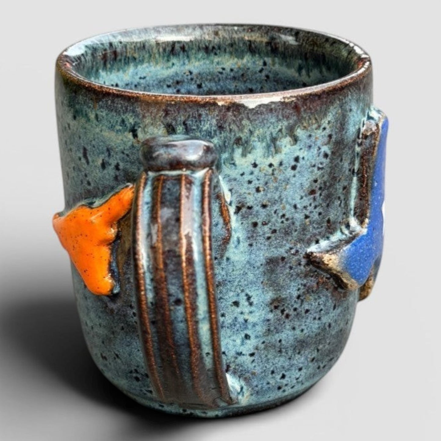 Rock N Art Shop handmade stoneware ceramic mug with speckled blue-green glaze and raised orange and blue state design.
