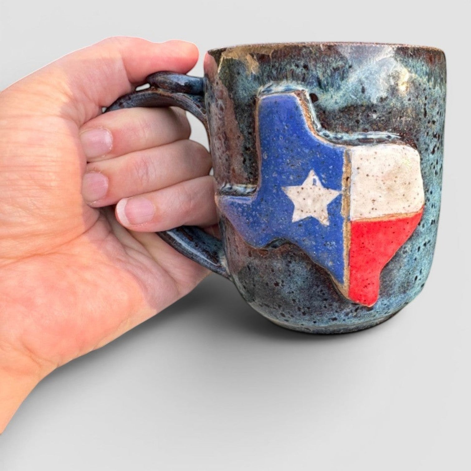 Rock N Art Shop handmade stoneware mug with a raised Texas flag design, speckled blue-green glaze.