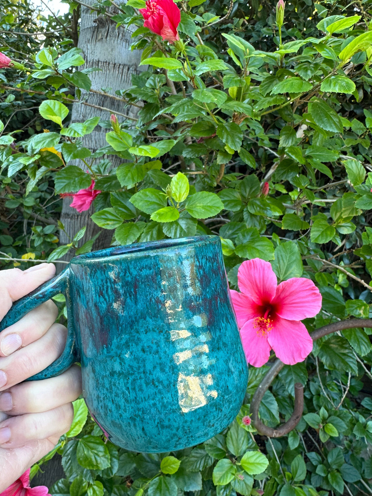 Rock N Art Shop handmade speckled stoneware coffee mug in turquoise blue and purple held against a hibiscus bush.
